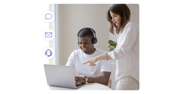 Agente di supporto sorridente con cuffie al laptop, un collega indica lo schermo; a sinistra, icone fluttuanti di chat, e-mail e chiamate vocali